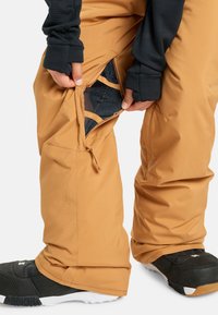 Hnedé snežné nohavice majú zipsové otváranie odhaľujúce sieťovanú podšívku, pričom na spodnej časti sú čierne lyžiarske topánky, čo zobrazuje elegantný a funkčný dizajn.
