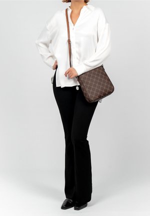 Femme portant un chemisier blanc et un pantalon noir, portant un sac à bandoulière marron à motifs avec une sangle réglable et une fermeture éclair ronde.