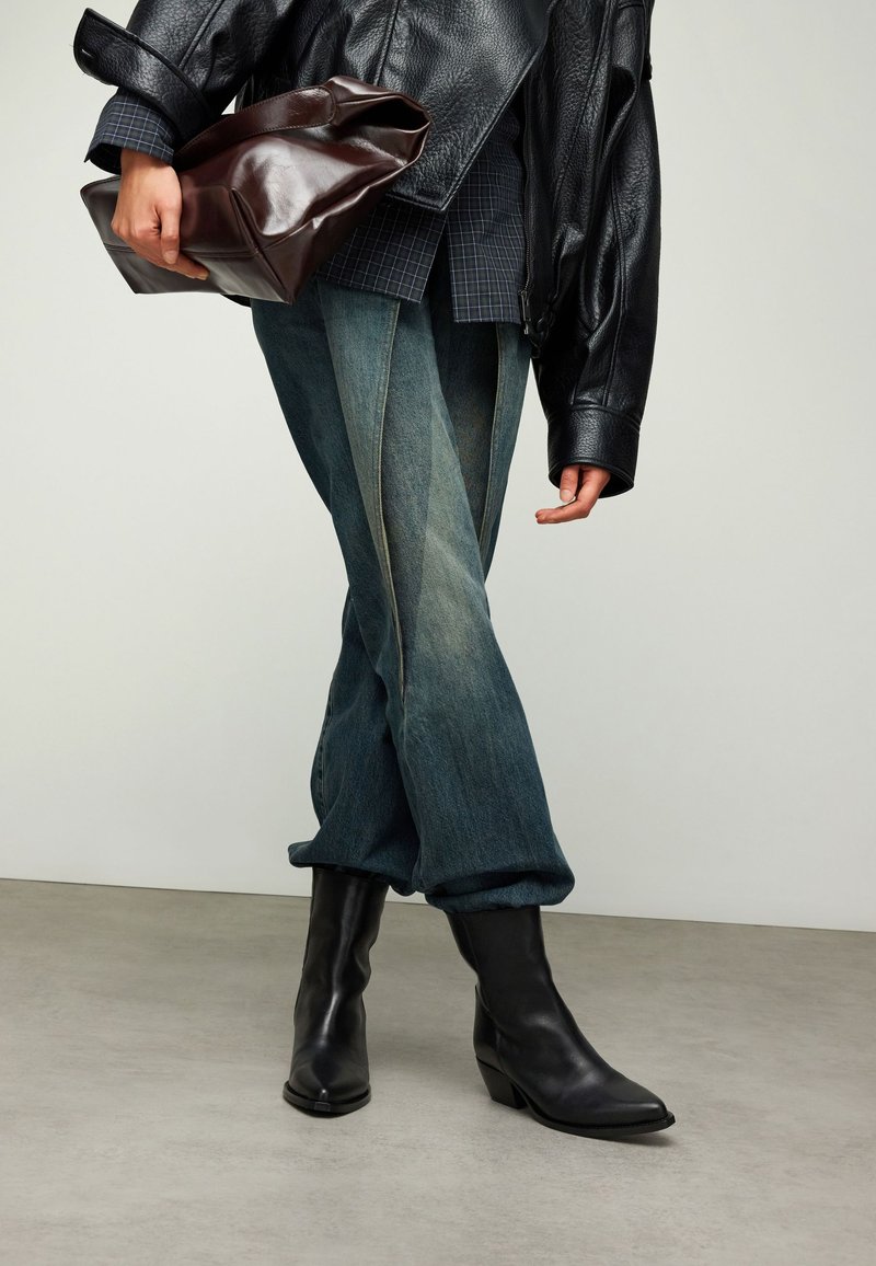 Veste en cuir noir, jean en denim bleu avec revers pliés, bottines noires et pochette en cuir marron. Des lignes épurées et des matériaux texturés.