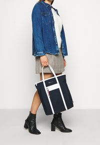 Marinblå tygväska med vita detaljer och ficka med dragkedja, tillverkad av hållbart material. Har två handtag och en strukturerad form.