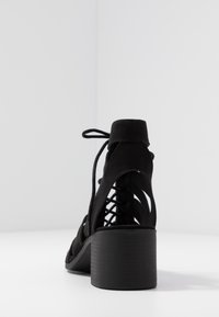 Czarne zamszowe sandały na obcasie z okrągłym noskiem, wiązaniem z tyłu, wycięciami i teksturowanym słupkiem.