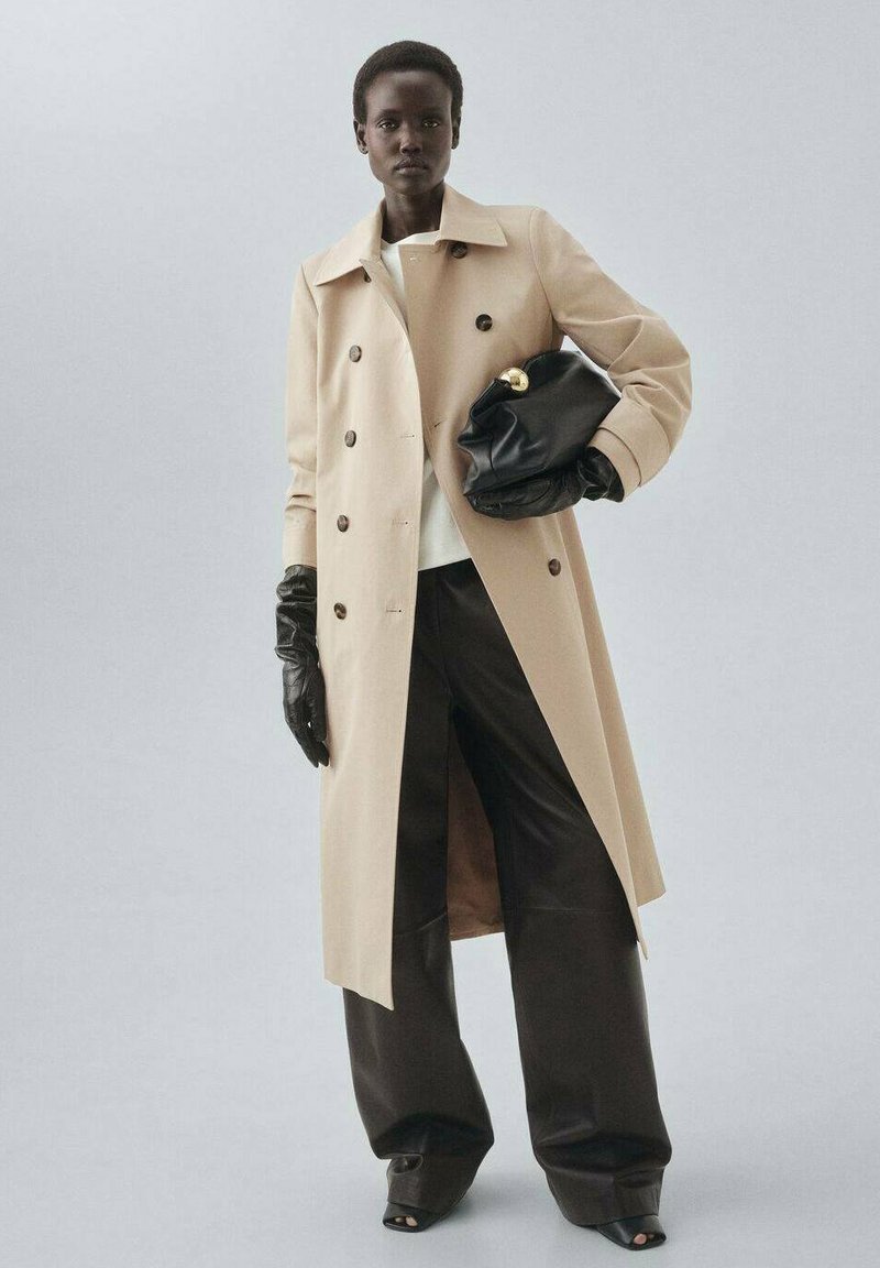 Beige trenchcoat, jossa on kaksirivinen malli, mustat nahkahanskat, ylisuuri musta laukku kultaisin yksityiskohdin ja tummat leveälahkeiset housut.