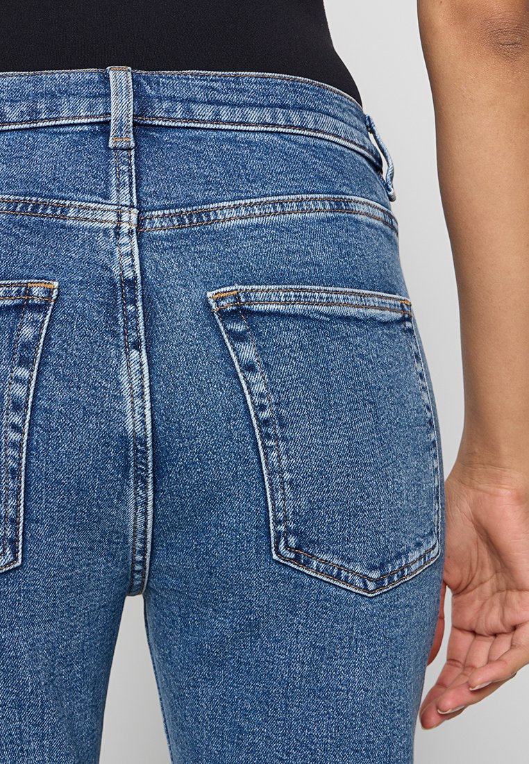 Blaue Denim-Jeans mit hoher Taille, ausgestattet mit zwei Gesäßtaschen und kontrastierenden Nähdetails. Die Textur wirkt grob und strapazierfähig.