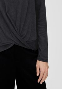 Rechte Hand einer Person, die ein dunkelgraues Langarmoberteil mit gedrehtem Frontdetail und schwarze Hose trägt.