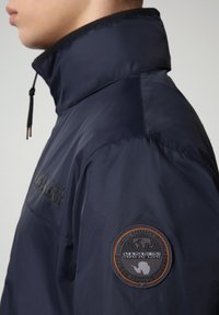 Giacca in nylon blu navy con colletto alto, dotata di una patch con logo sulla manica sinistra e una texture liscia e leggera.