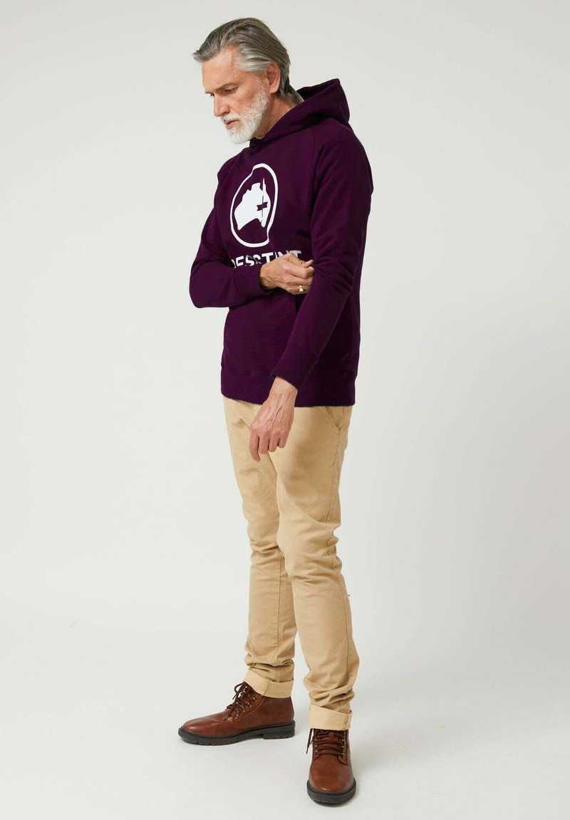 Sudadera con capucha morada con un logo blanco, pantalones beige y botas marrones con cordones. La sudadera tiene un bolsillo frontal y puños acanalados.