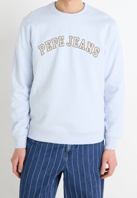 Sudadera azul claro con puños y dobladillo acanalados, con texto "PEPE JEANS" en forma de arco, mostrado en color beige. Combinada con pantalones azules a rayas.