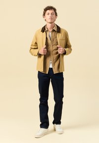Giovane uomo con capelli ricci sistema la giacca beige sopra una camicia marrone, jeans scuri e sneakers bianche, in piedi davanti a uno sfondo beige chiaro.