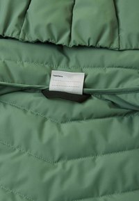 Wewnętrzna strona zielonej kurtki wypełnionej, z krzyżowym pikowaniem. Zawiera metkę z napisem "Byłem tutaj:". Gładka tekstura.