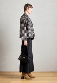 Veste à motifs noir et fauve, robe noire jusqu'aux chevilles, bottines à talons beiges, tenant un sac à main noir avec un accent doré.