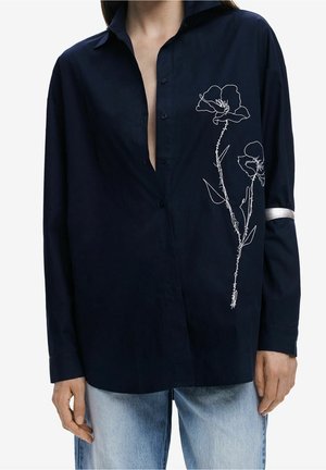 Person iført en mørk marineblå skjorte med knapper og hvid blomstret linjekunst på brystet samt et sølvbånd på højre ærmekant.