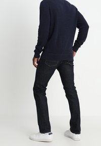 Jeans in denim blu scuro con una vestibilità slim, abbinati a un maglione blu navy. I jeans presentano tasche posteriori e una texture liscia; sneakers bianche completano il look.