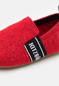 Une chaussure rouge en feutre, à enfiler, dotée d'un bandeau élastique noir avec un texte blanc et d'un dessus texturé. Semelle en caoutchouc avec un design minimaliste.