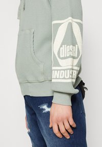 Grön texturerad hoodie med en vit logotyp på ärmen. Har en framfick och ribbade muddar, ihop med slitna blå jeans.