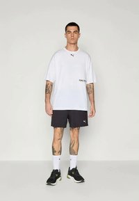 Bijela oversized majica s crnim PUMA logotipom, uparena s crnim šortsevima. Bijele čarape i crne sportske cipele upotpunjuju odjevnu kombinaciju.