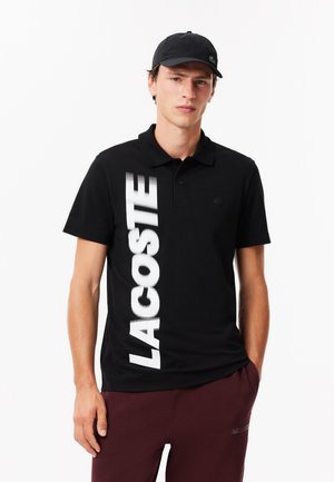 Czarny polo wykonany z bawełny, z pionowym białym logo "LACOSTE" po lewej stronie oraz małym zielonym emblematem krokodyla na piersi.