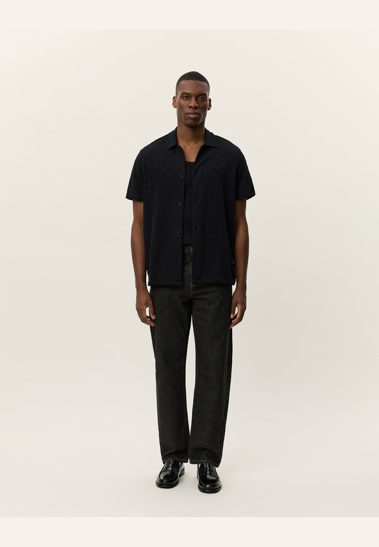 Camisa preta de mangas curtas com padrões texturizados, frente com botões, combinada com calças de ganga escuras e sapatos pretos. Modelo está de pé contra um fundo neutro.