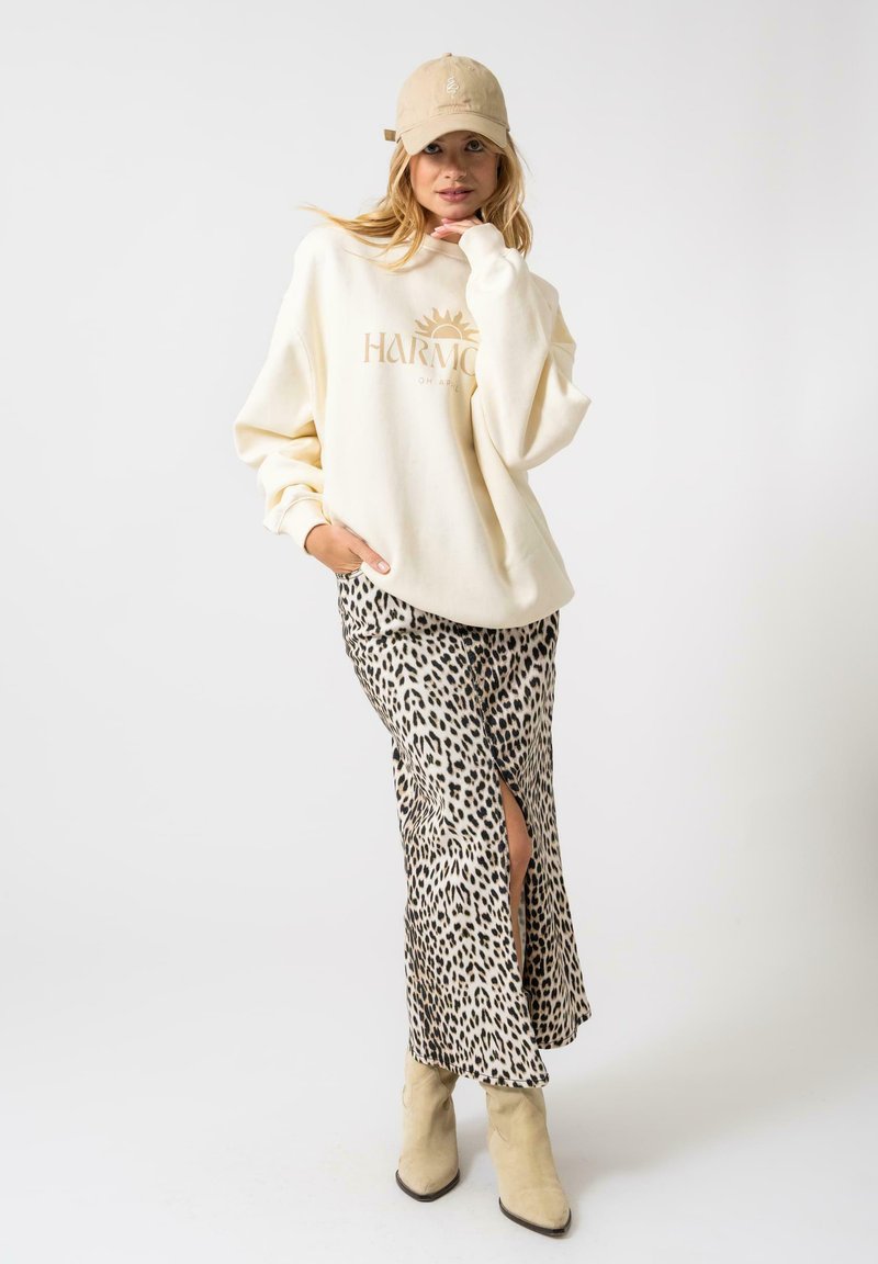 Pulover crem cu logo-ul "HARMONY", asociat cu o fustă cu imprimeu leopard și o fanta laterală. Modelul poartă o șapcă bej și botine.