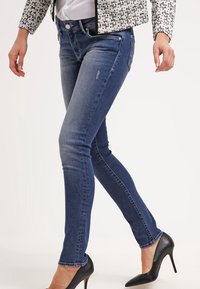 Vaqueros ajustados azules de mezclilla con un patrón desgastado, cinco bolsillos y cierre de botón, combinados con tacones altos negros.