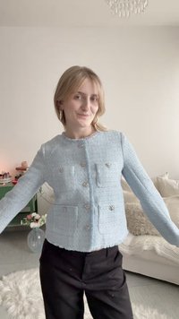 Νέα γυναίκα που φοράει ανοιχτό μπλε πλεκτή ζακέτα με κουμπιά και μαύρο παντελόνι, στέκεται σε καθιστικό με καναπέ και λουλούδια στο φόντο.