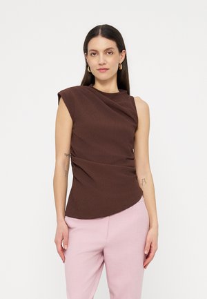 Kobieta o długich ciemnych włosach, ubrana w brązowy top na ramiączkach i jasnoróżowe spodnie, stoi na tle jednolitego białego tła.