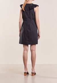 Černé šaty bez rukávů s volánkovým detailem na rameni a rovný střihem, vyrobené z hladké látky, zobrazené zezadu. Hnědé boty na širokém podpatku.