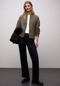 Chaqueta bomber a rayas beige con puños acanalados, camiseta blanca, vaqueros azul oscuro y botas negras hasta el tobillo. Modelo sosteniendo una chaqueta acolchada negra.