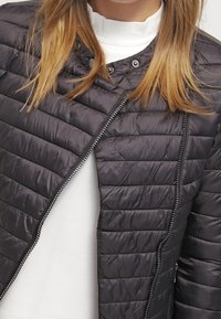 Chaqueta negra acolchada con cremallera frontal, fabricada con material ligero. Presenta un cuello alto y una textura suave y brillante.