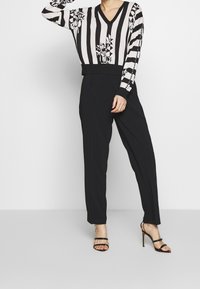Czarny i biały sweter w paski z kwiatowymi akcentami, dekold w serek, zestawiony z długimi czarnymi spodniami i czarnymi sandałami na wysokim obcasie.