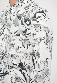 Camicia bianca con motivi floreali ondulati in nero e grigio, dotata di colletto classico e dettagli sui polsini. Logo visibile sulla manica.