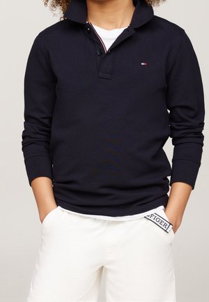 Niño con polo de manga larga azul marino con cuello y pantalones cortos blancos, manos en los bolsillos, pequeño logo de la marca en el pecho.