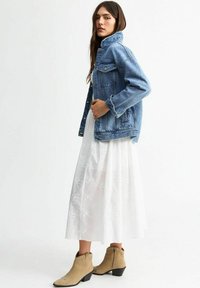 Giacca di jeans con taglio classico, lavaggio chiaro, dotata di tasche frontali e chiusura con bottoni, abbinata a un vestito midi bianco ricamato e stivaletti beige alla caviglia.