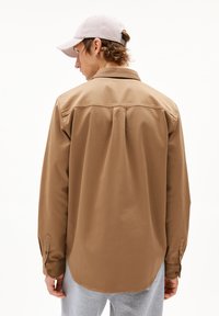 Camicia marrone a maniche lunghe con polsino con bottoni, dettaglio a piega sul retro e orlo arrotondato, abbinata a un berretto rosa chiaro e pantaloni grigi.