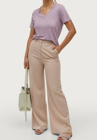 Svetle fialové tričko s výstřihem do V v kombinaci s béžovými kalhotami s širokými nohavicemi. Oblečení doplňuje měkká zelená kabelka a světle šedé sandály.