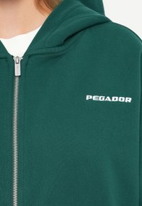Felpa verde con zip realizzata in tessuto testurizzato. Presenta un cappuccio e il logo ricamato "PEGADOR" in bianco sul petto. Dettaglio della zip argento.
