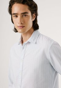 Gaiši zils krekls ar plānām baltām svītrām, garām piedurknēm un strukturētu apkakli. Izgatavots no mīkstas, teksturētas auduma.