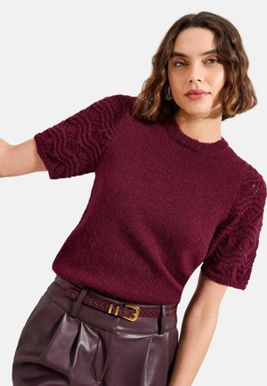 Nainen, jolla on päällään viininpunainen teksturoitu lyhythihainen neule ja vyötäröä korostavat viininpunaiset nahkahousut vyöllä, poseeraa yksinkertaista taustaa vasten.