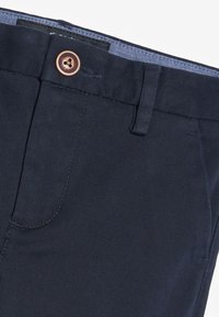 Partie supérieure avant d’un pantalon bleu marine montrant une fermeture à bouton, une passoire de ceinture et une partie de la poche avec des détails de coutures visibles.