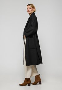 Abrigo largo negro con mangas anchas, bolsillos laterales y un cuello sencillo, combinado con pantalones color crema y botines marrones. Textura de tela suave.