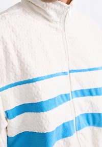 Fleece bunda s vysokým límcem, s texturovaným krémovým tělem a dvěma horizontálními modr ými pásky. Zapínání na zip přední.