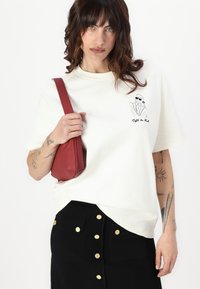 Cream t-shirt with short sleeves and graphic print on the chest, paired with a black skirt featuring gold buttons and pockets, and a red bag.