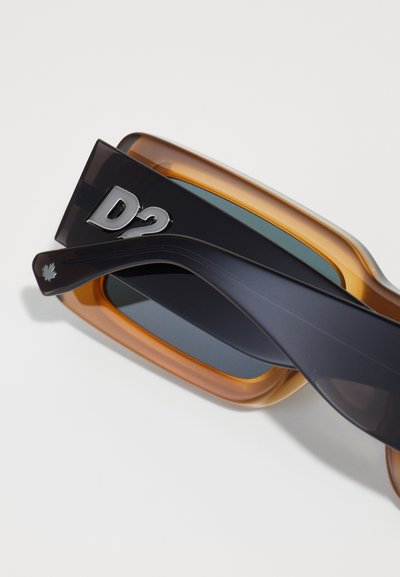 Sunglasses with a clear and dark navy frame, brown tinted lenses, and a silver D2 logo on the side. Smooth texture and oversized rectangular shape.