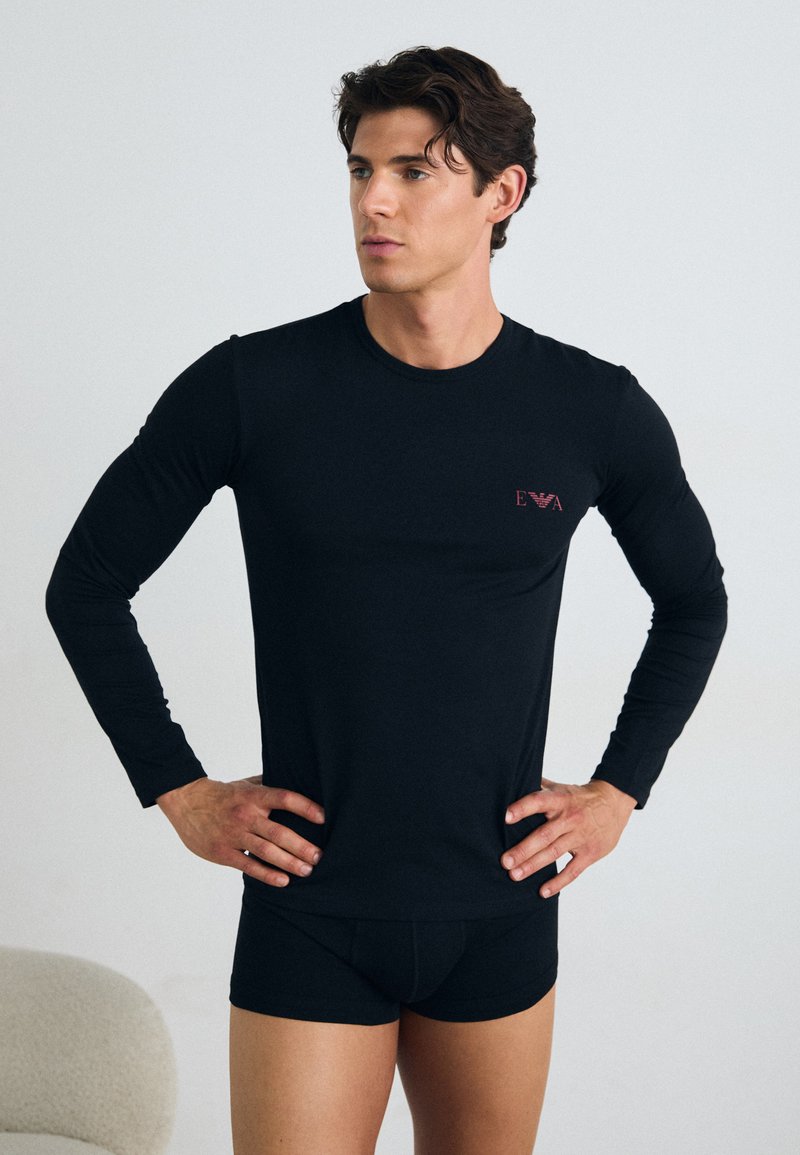 Chemise noire à manches longues avec un design ajusté et un petit logo rouge sur la poitrine, accompagnée de shorts noirs ajustés. Texture lisse.