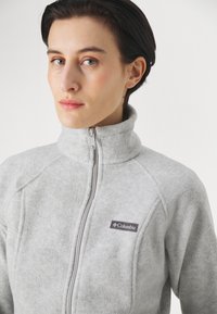 Vaaleanharmaa fleece-takki, jossa on korkea kaulus, täysi vetoketju ja raglan-hihat. Takissa on pieni tummanharmaa Columbia-logo rinnassa.