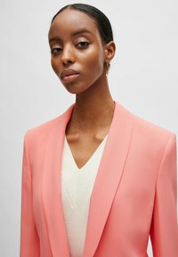 Pink blazer with a smooth texture and shawl collar, layered over a cream v-neck sweater with subtle ribbing detail.