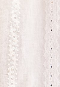 Tissu blanc avec deux bordures de dentelle brodée verticales ; l'une avec de petits motifs floraux, l'autre avec des bords festonnés et de minuscules points noirs.