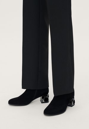 Stivaletti neri in suede con punta quadrata e tacco decorativo geometrico nero, abbinati a pantaloni neri su misura.