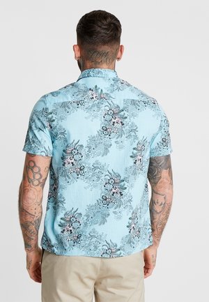 Mand med tatoveringer iført en lyseblå kortærmet blomsterprintet skjorte og beige bukser, vist bagfra mod en ensfarvet baggrund.