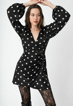 Femme portant une robe portefeuille noire à pois avec des manches ballon, des collants en dentelle noire et des bottes noires, posant les mains au-dessus de la tête.