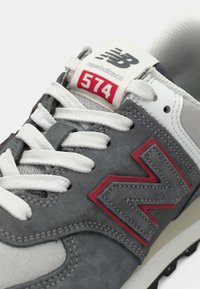 Grå ruskind sportssko med hvide mesh-detaljer, rødt branding og hvide snørebånd. Har en polstret sål for komfort. Model 574.